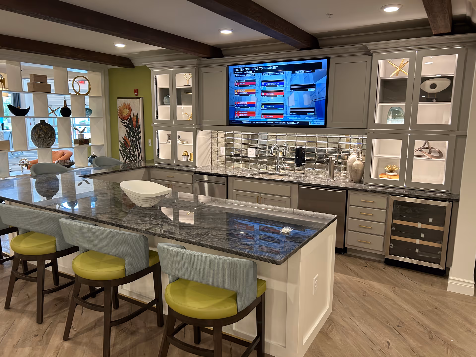 Modern kitchen area in a senior living facility with a large island countertop featuring four chairs with green cushions. The kitchen has white cabinets with glass display sections, a mirrored backsplash, and built-in appliances including a dishwasher and wine cooler. A flat-screen TV is mounted above the sink area, and decorative items are displayed on shelves and inside cabinets. The flooring is wood-style, and there are exposed wooden beams on the ceiling.