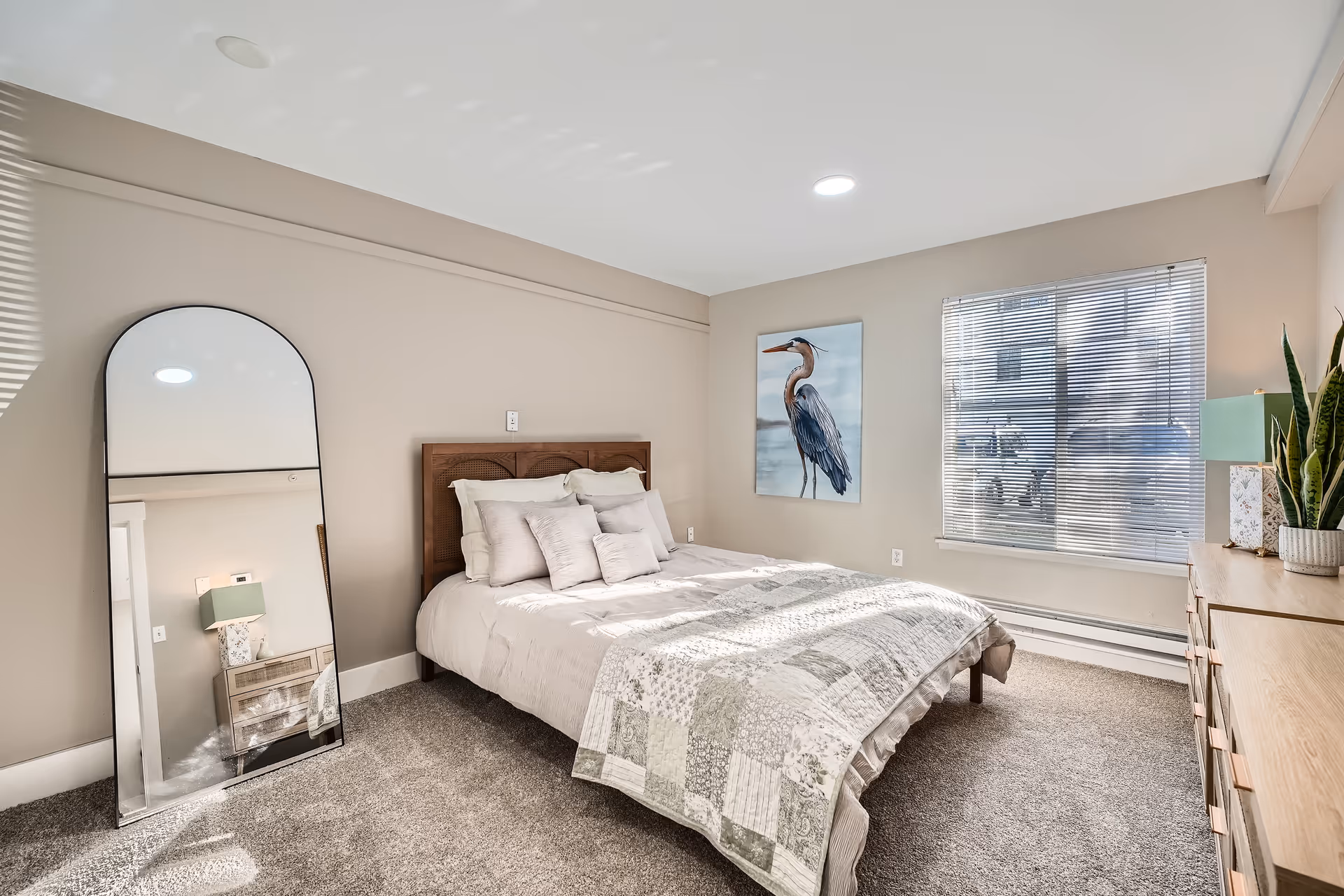 Bright bedroom at Farrington Court with a quilted bed, arched mirror, dresser, and window blinds.
