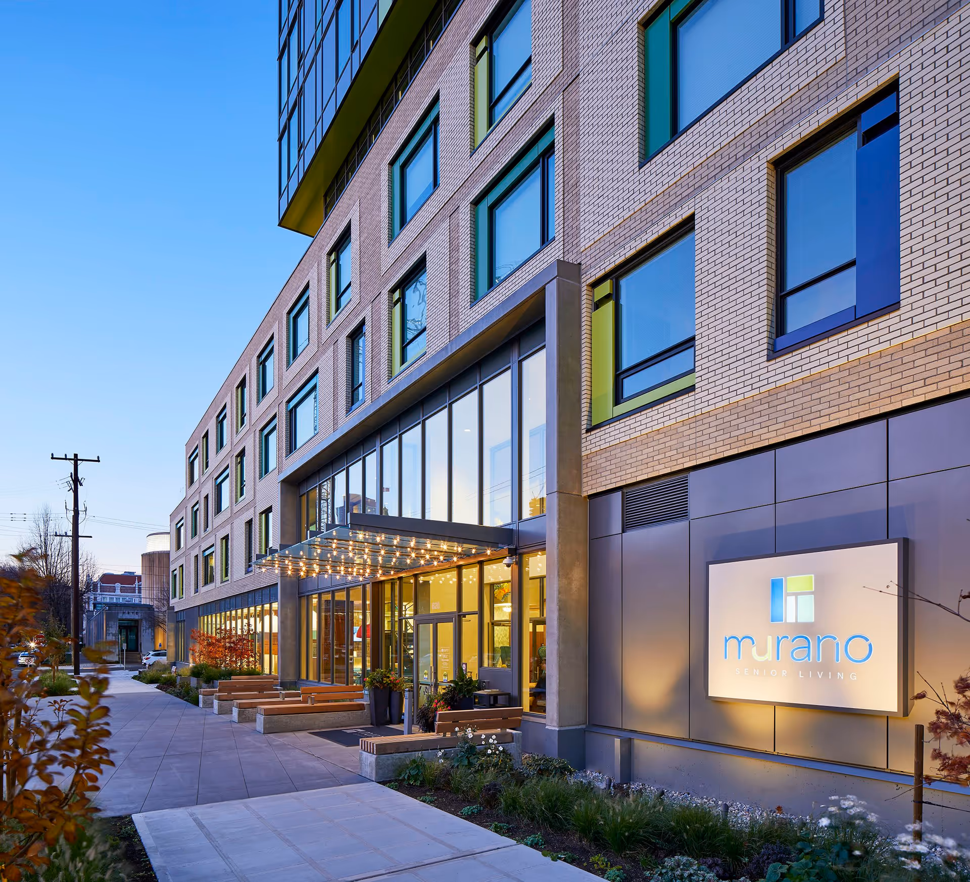 Exterior street-level view of the modern Murano Senior Living building showing large windows, an illuminated entrance canopy, and the Murano sign.