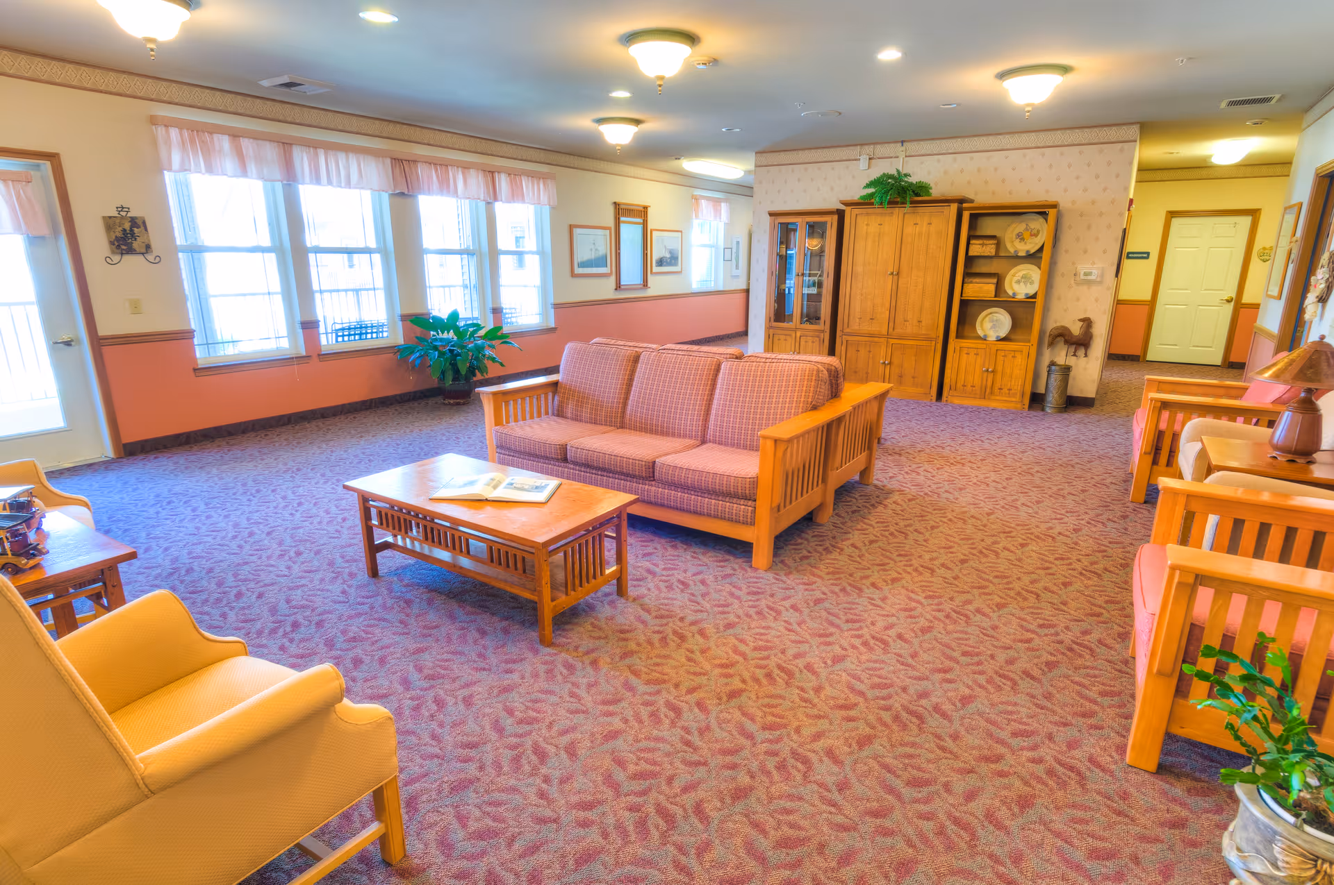 A spacious living room area in a senior living facility with patterned carpet, wooden furniture including a sofa, armchairs, coffee table, and cabinets. The room is well-lit with ceiling lights and natural light from large windows with sheer curtains. There are decorative plants and framed pictures on the walls.