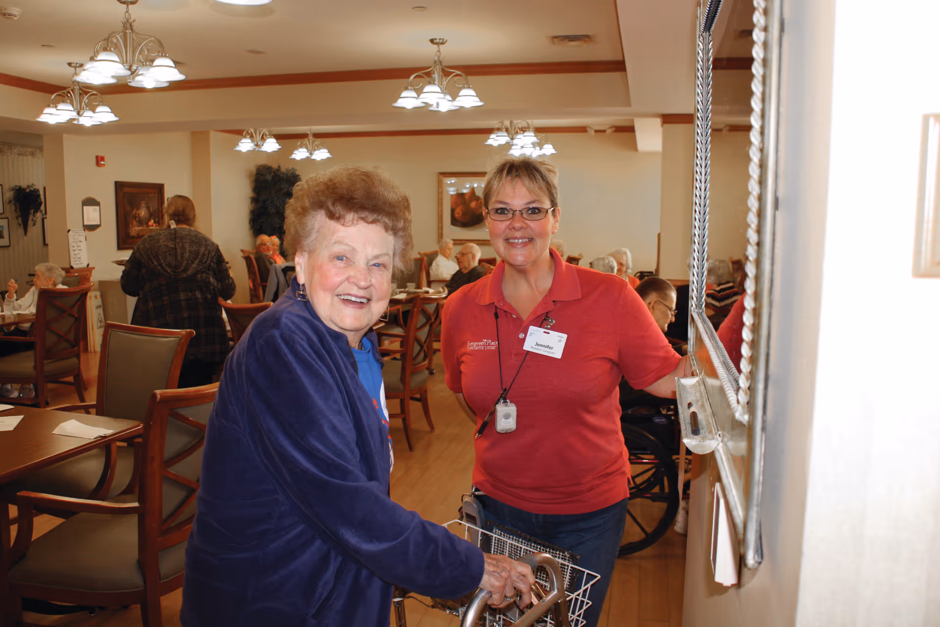 An elderly woman with a walker smiling at the camera, accompanied by a female staff member wearing a red shirt and name tag, inside a dining room with several other elderly residents seated at tables in the background.