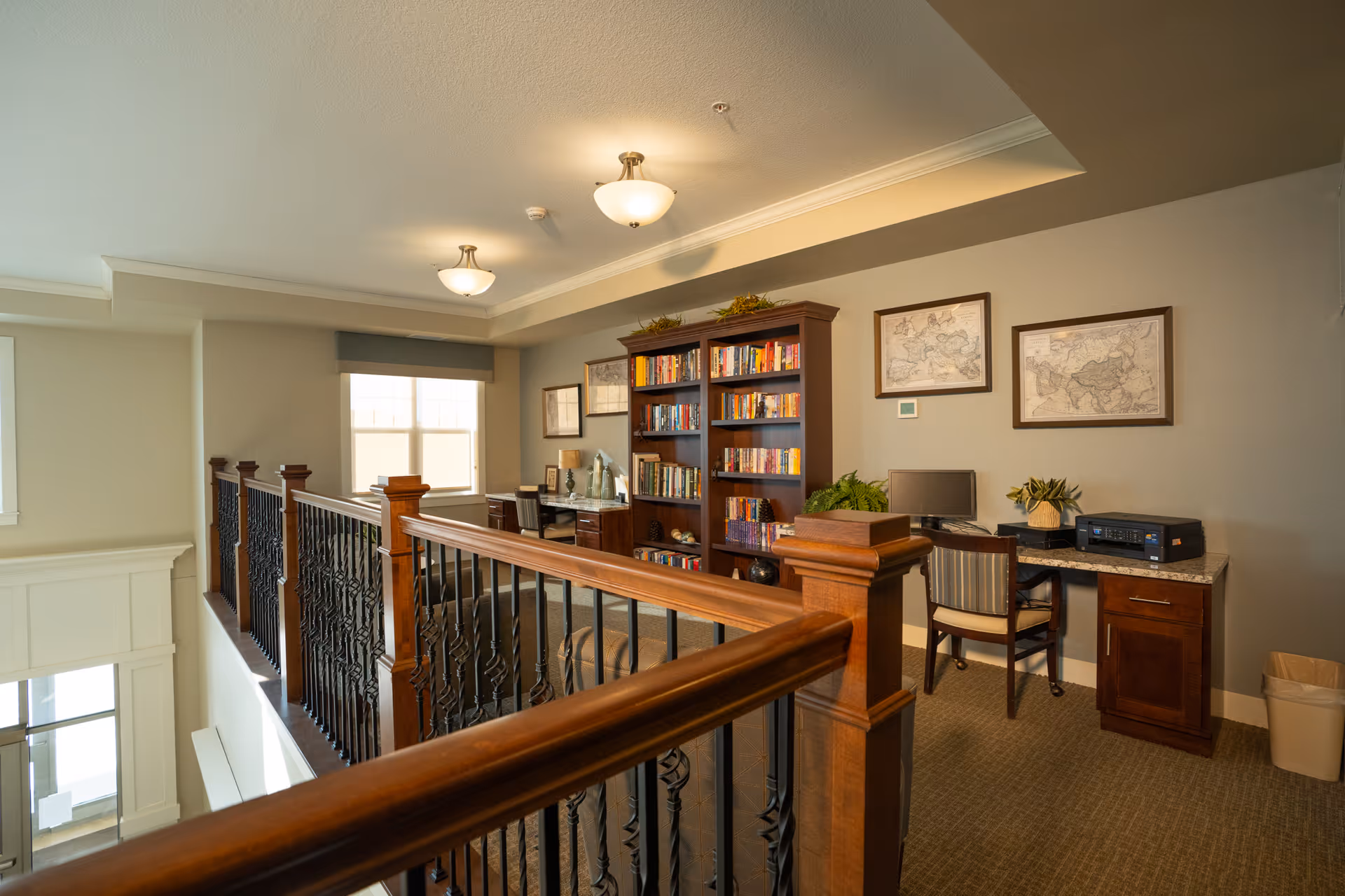 Interior view of a senior living facility featuring a small office area with two desks, chairs, a computer, a printer, and a bookshelf filled with books. The space has beige walls, carpeted floors, decorative framed maps on the wall, and a wooden railing with black metal balusters overlooking a lower level.