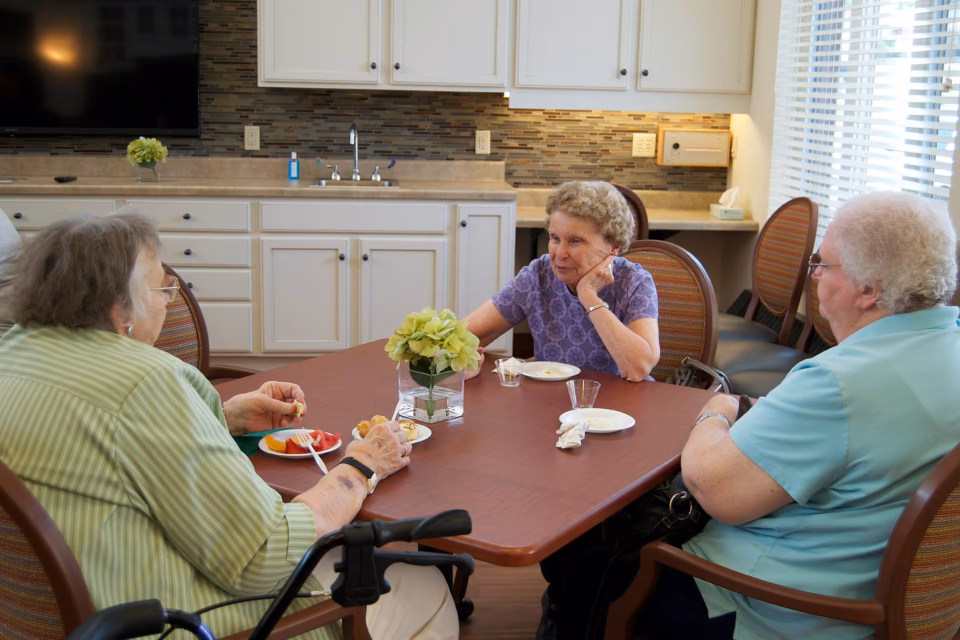 Three elderly women sitting around a wooden table in a well-lit room with a kitchen area in the background. They are engaged in conversation and have plates with food in front of them. The room has white cabinets, a tiled backsplash, and a window with blinds letting in natural light.