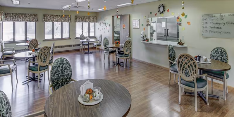 Cozy senior living dining room with round tables and green upholstered chairs, decorations on walls.
