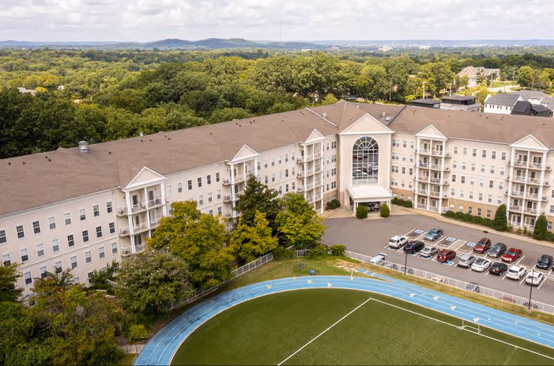 Aerial view of a large senior living community building with multiple floors and balconies, surrounded by trees and greenery. In front of the building is a parking lot with several cars parked, and a blue running track encircling a green sports field.