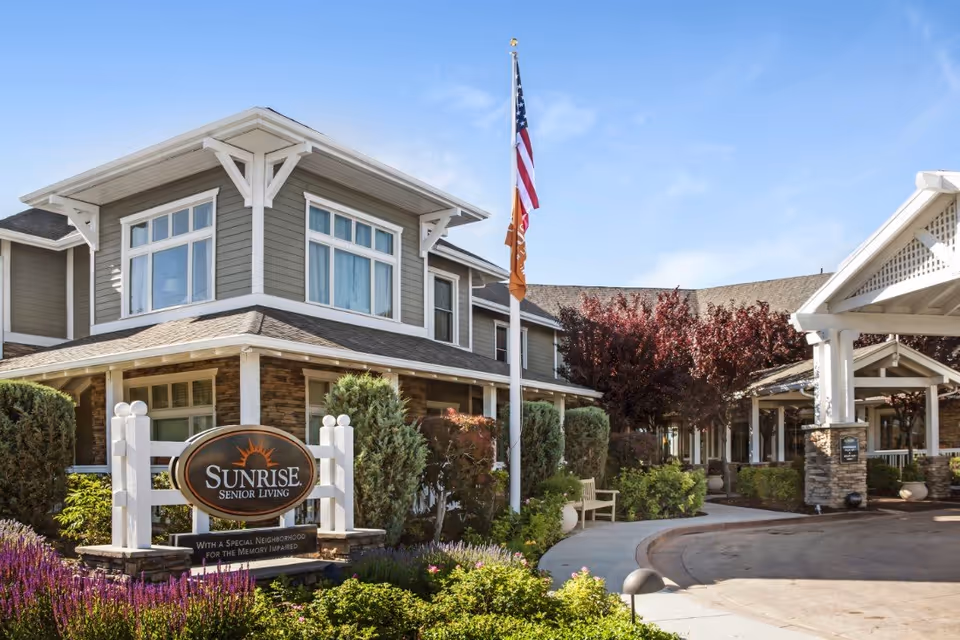 Front entrance of the Sunrise Senior Living facility with a sign, flagpole, landscaped beds, and a covered drive-up.