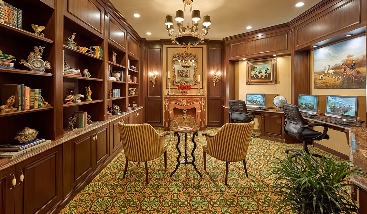 A cozy, elegant room with wooden bookshelves filled with books and decorative items on the left, two striped armchairs facing a small round table in the center, and a fireplace with a decorative mirror above it on the far wall. On the right side, there are two desks with computers and office chairs, along with framed paintings on the wall. The room has a patterned carpet and warm lighting from a chandelier and wall sconces.
