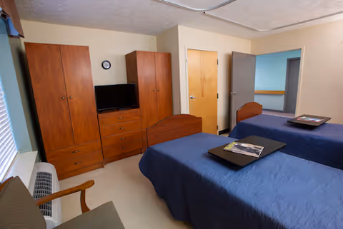 Two single beds with blue bedspreads in a shared senior living bedroom with wardrobes and TV