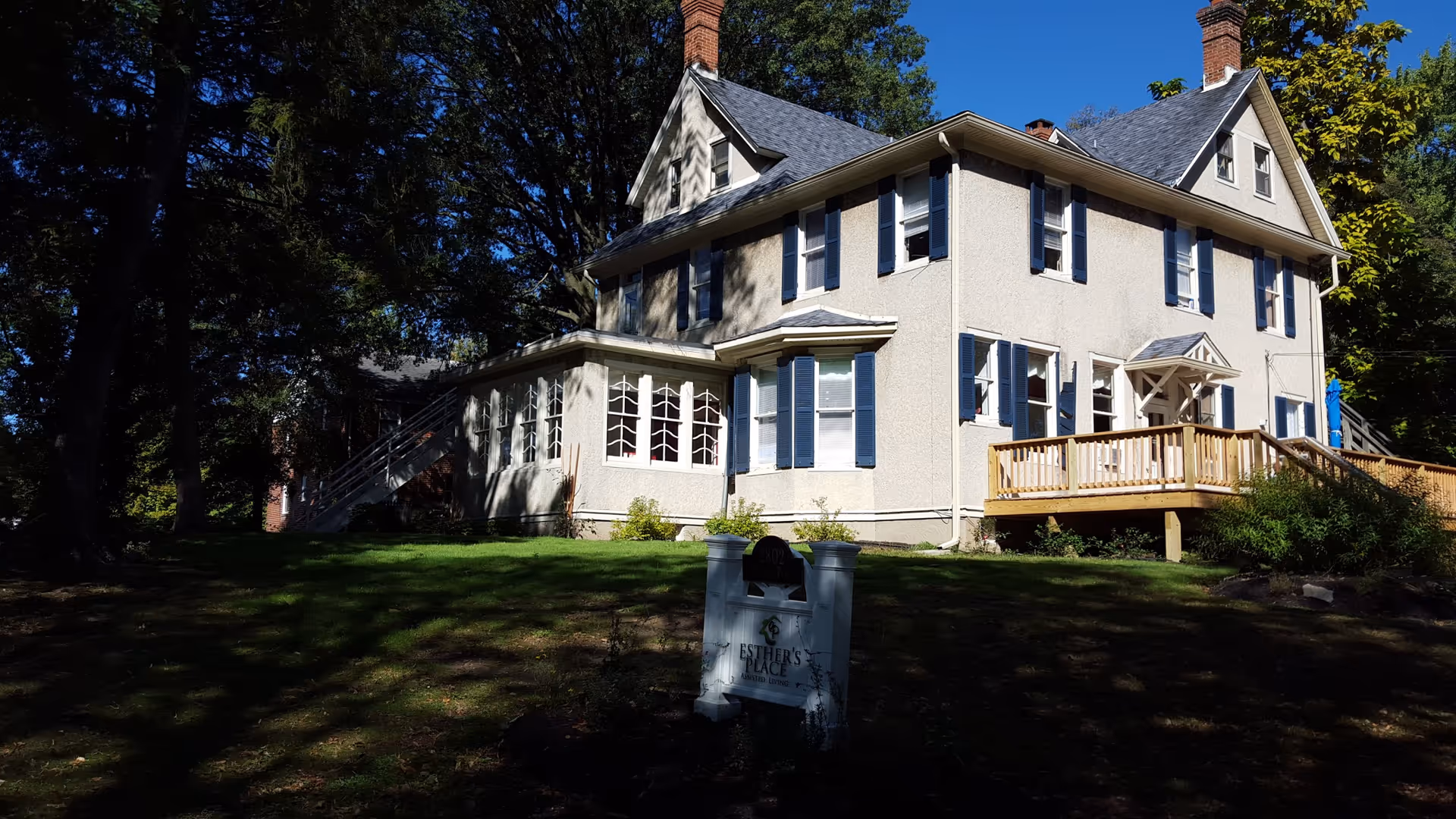 A large two-story house with beige exterior walls, blue shutters, and a wooden deck at the front. The house is surrounded by trees and grass, with a sign in the front yard that reads 'Esther's Place'.