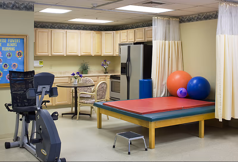 Interior therapy room with an exercise bike, a padded treatment table and mats, colorful exercise balls, and a small kitchenette with seating.