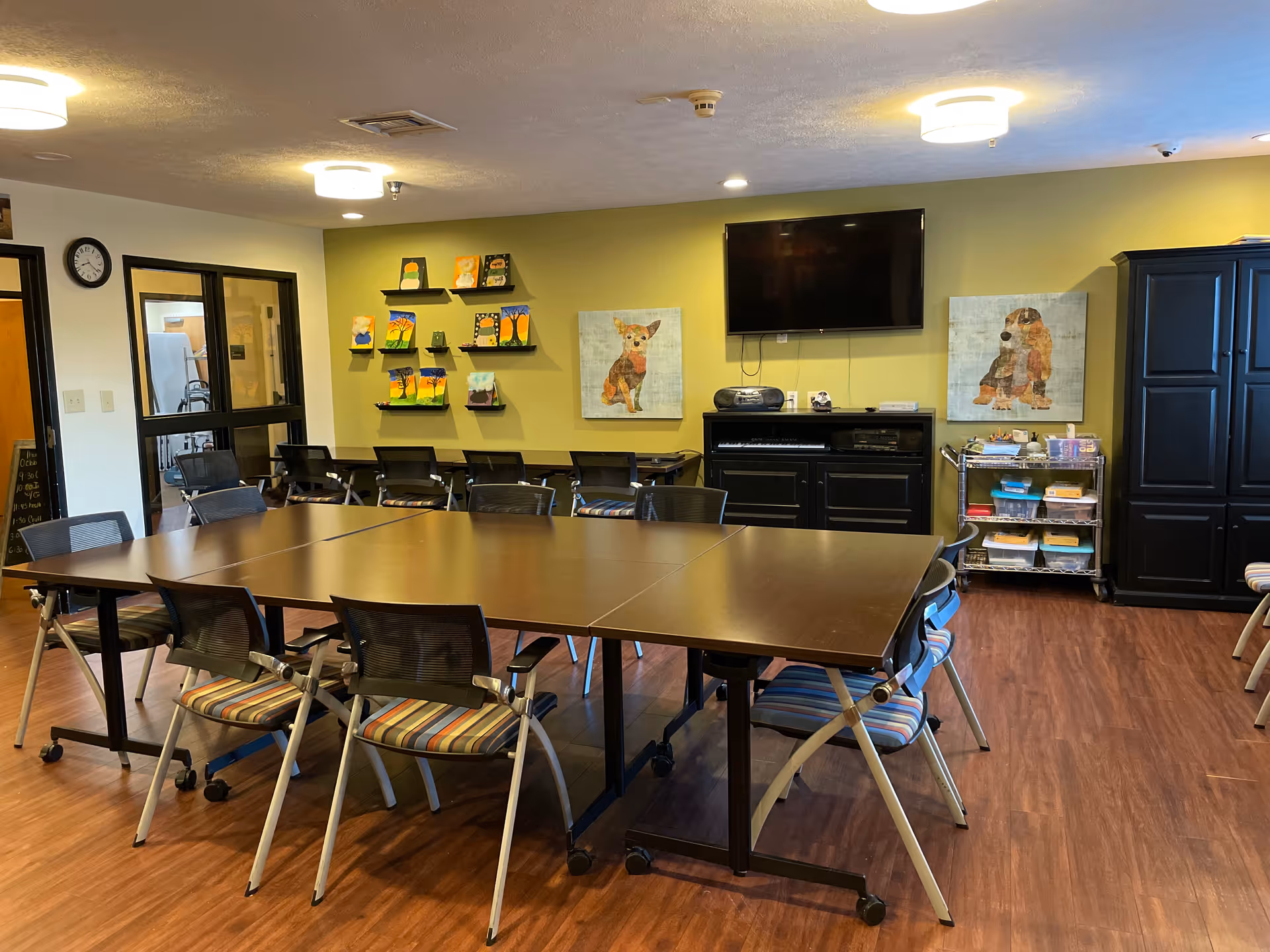 A well-lit activity room with a large rectangular table surrounded by chairs with striped cushions. The walls are painted yellow and decorated with colorful paintings of dogs and small shelves holding art supplies. A flat-screen TV is mounted on the wall above a black cabinet, and a rolling cart with various materials is positioned nearby. The floor is wood laminate, and there is a clock on the wall near a glass door.
