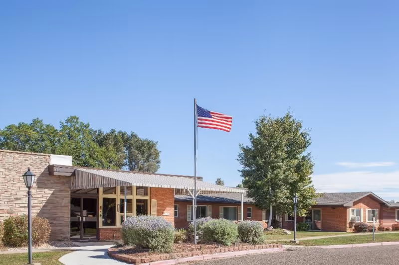 Front exterior of the Atlas Post Acute building showing its entrance, landscaped shrubs, and an American flag against a clear blue sky.