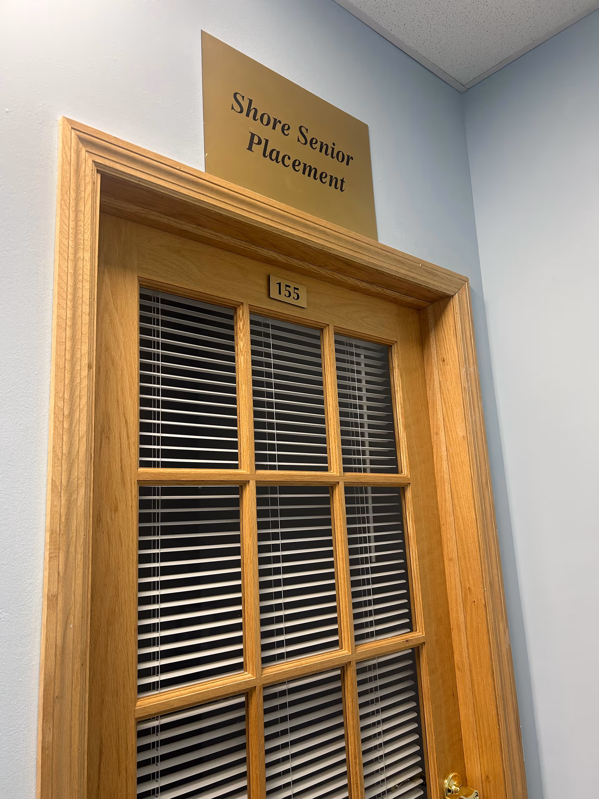 Wood-framed door with blinds and a Shore Senior Placement sign in a hallway.