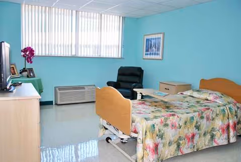 A resident room in a senior care facility with a floral bedspread, chair, and window blinds.