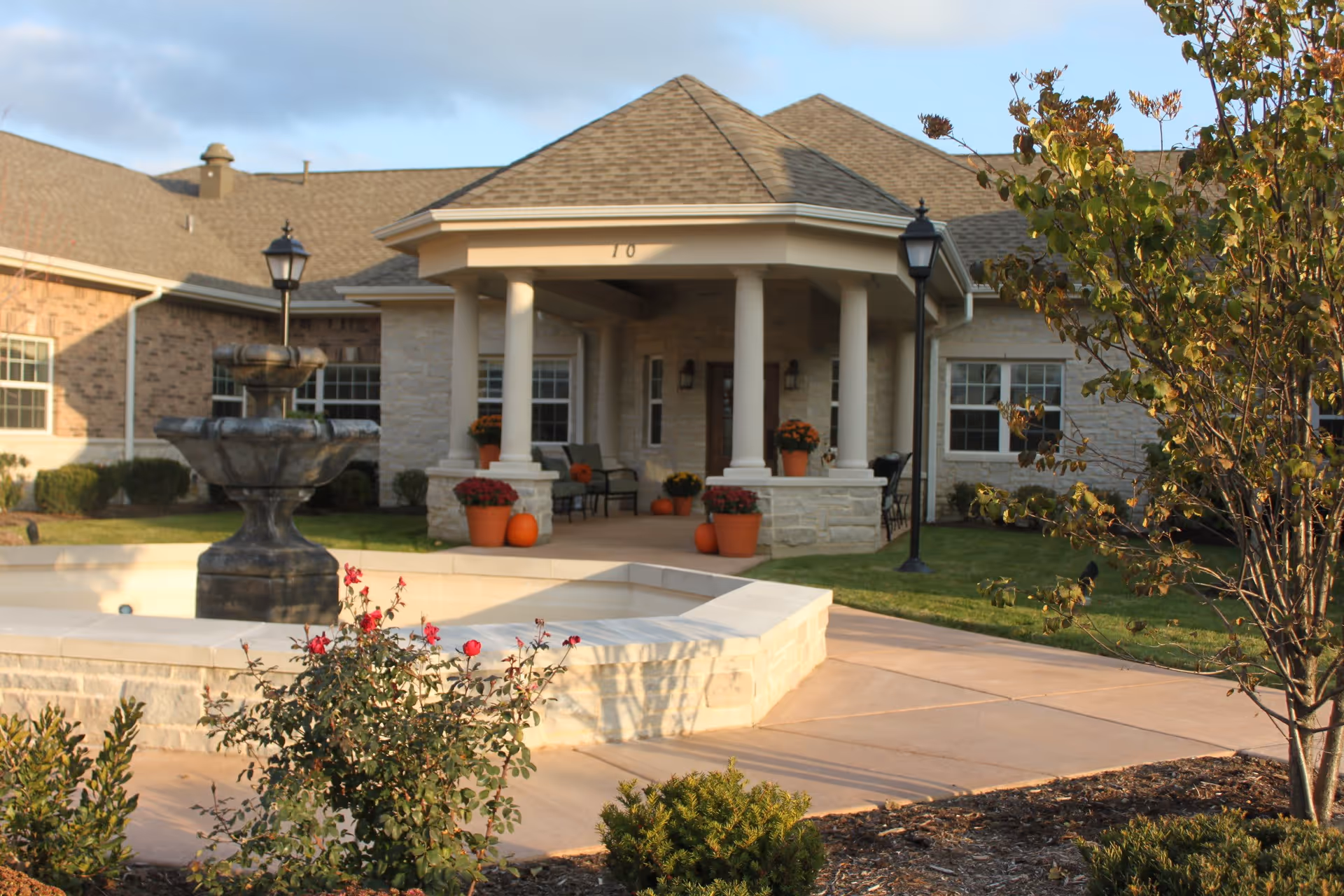 Front entrance of a senior living facility with a covered portico, stone fountain, potted pumpkins and landscaped walkways.