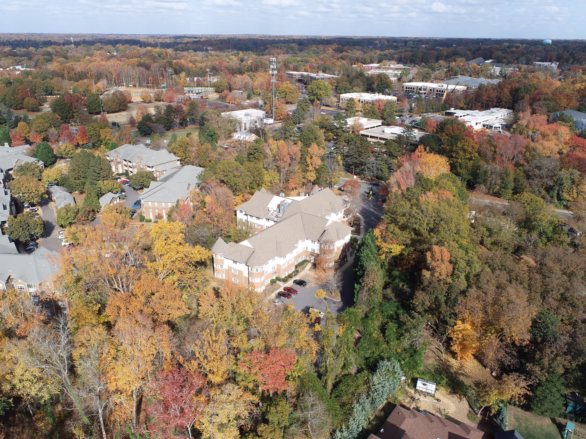 Aerial view of a senior living facility surrounded by dense trees with autumn foliage in various shades of yellow, orange, red, and green. The facility consists of multiple multi-story buildings with beige roofs and brick walls. Nearby are other residential and commercial buildings, parking lots, and a water tower in the distance under a partly cloudy sky.
