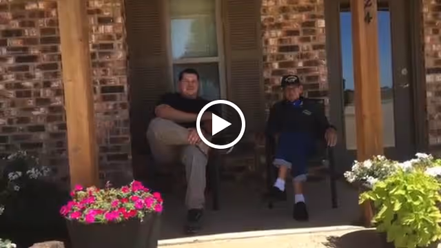 Two men sitting on chairs on a covered porch in front of a brick building with a wooden post and flower pots with pink and white flowers.