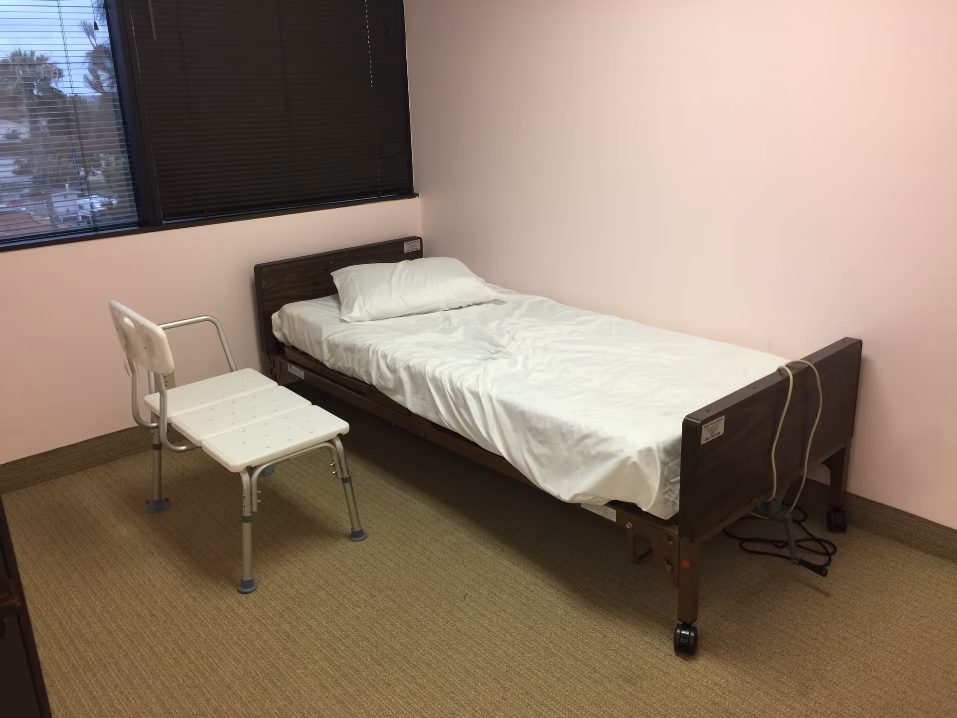 A single hospital-style bed and chair in a pink-walled senior living room.