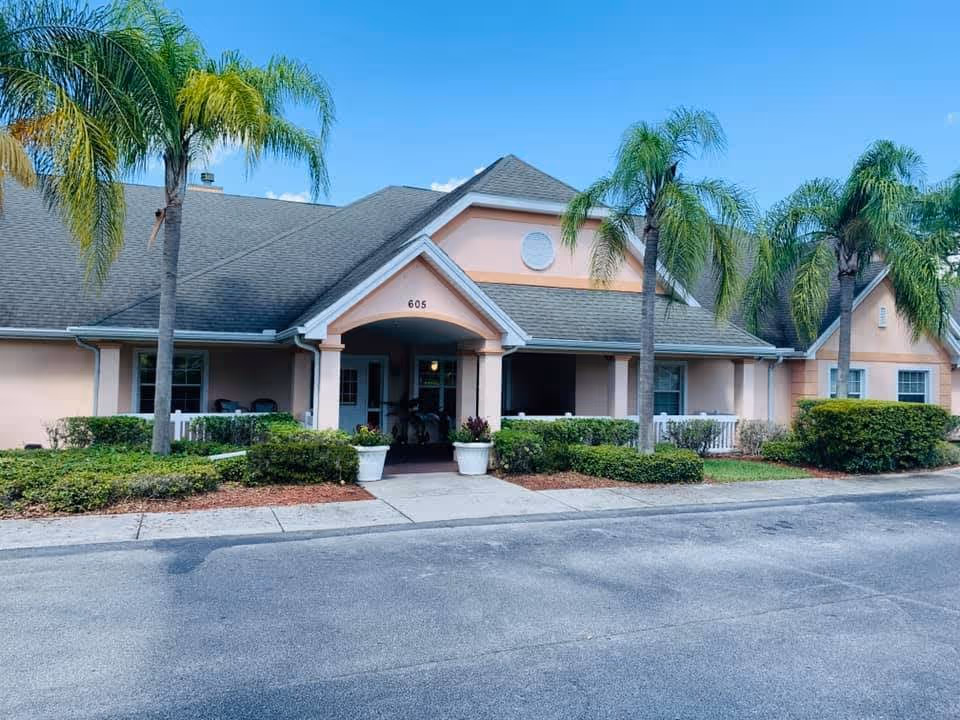 Exterior view of a single-story building with a gray shingled roof and peach-colored walls, surrounded by palm trees and green shrubs. The building has a covered entrance with the number 605 above it and two large white planters with flowers on either side of the walkway leading to the door. The sky is clear and blue.