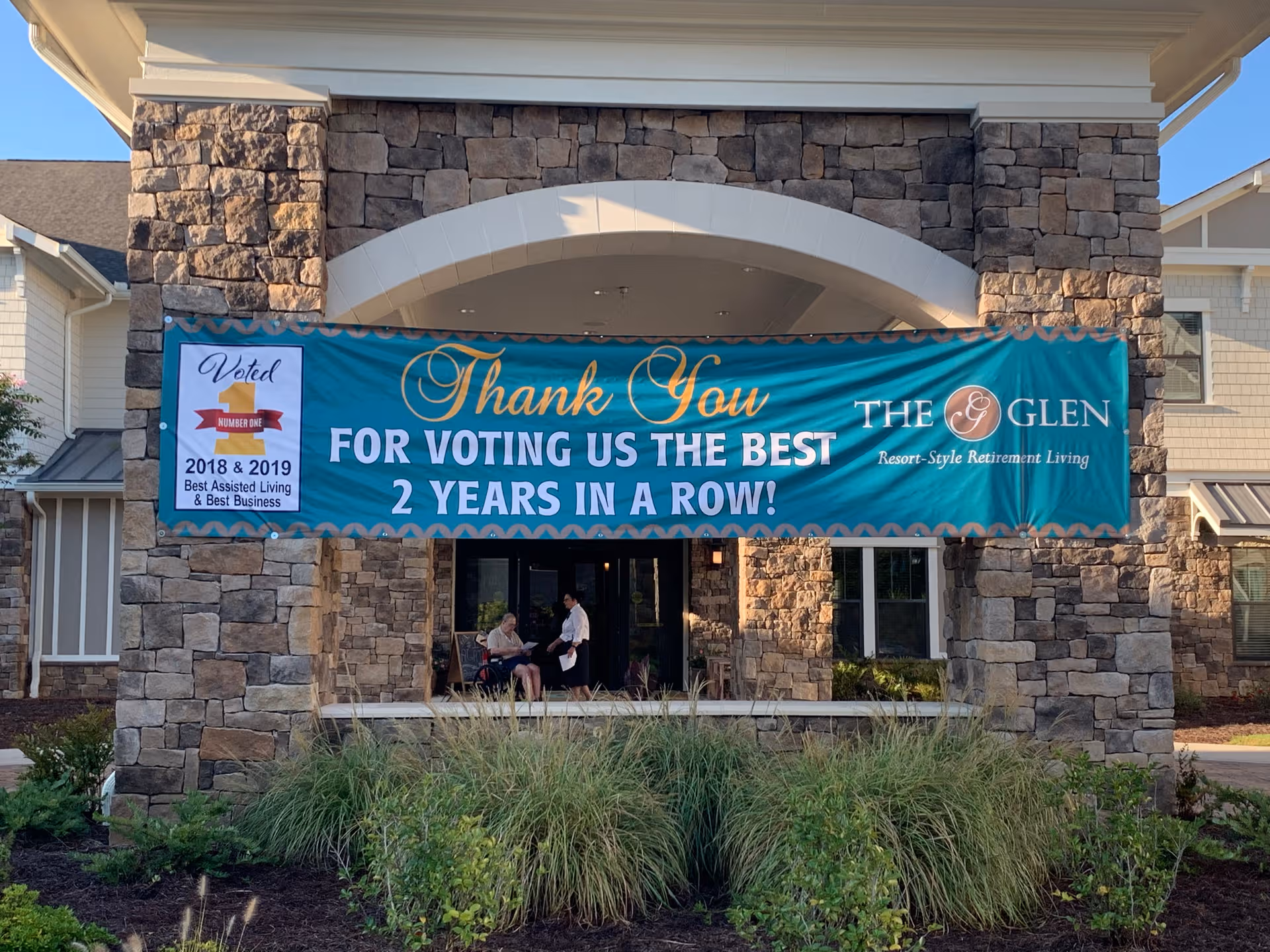 Stone-front entrance with banner at The Glen at Lake Oconee Village and two people near doors