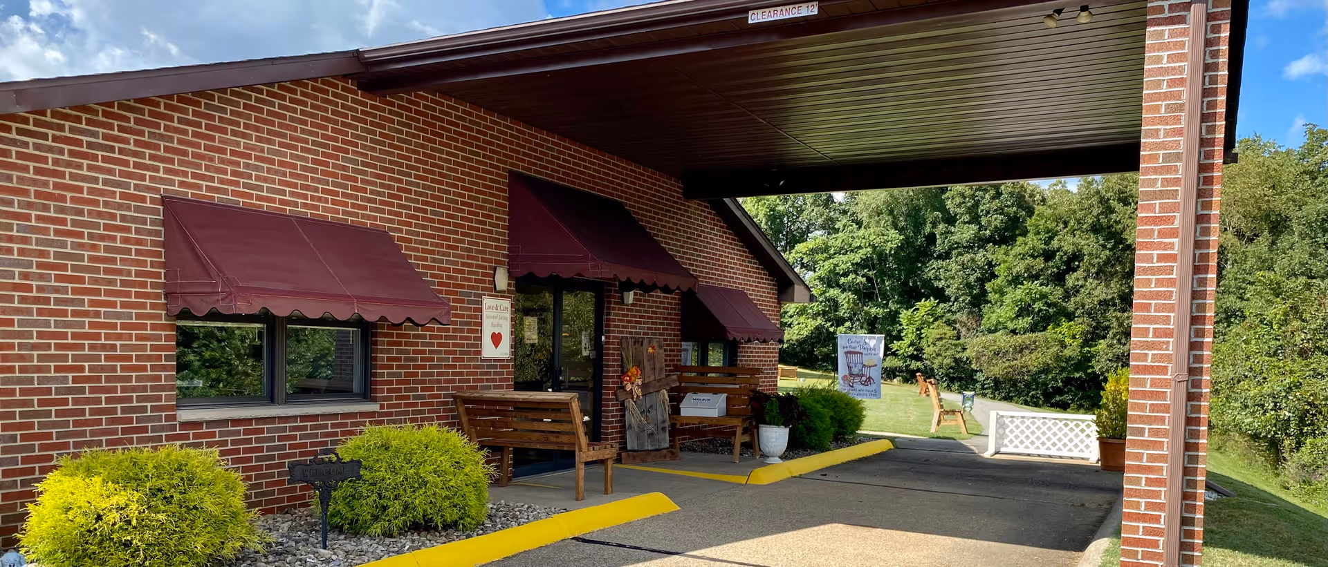 Brick single-story building entrance with a covered drive-through canopy, maroon awnings, benches, and shrubs.