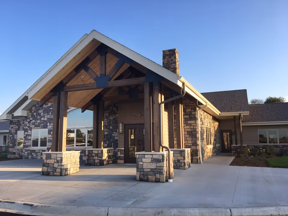 Exterior view of The Legacy of DeForest facility showing a stone and wood entrance with a covered porch supported by wooden beams, a chimney, and a clear blue sky.