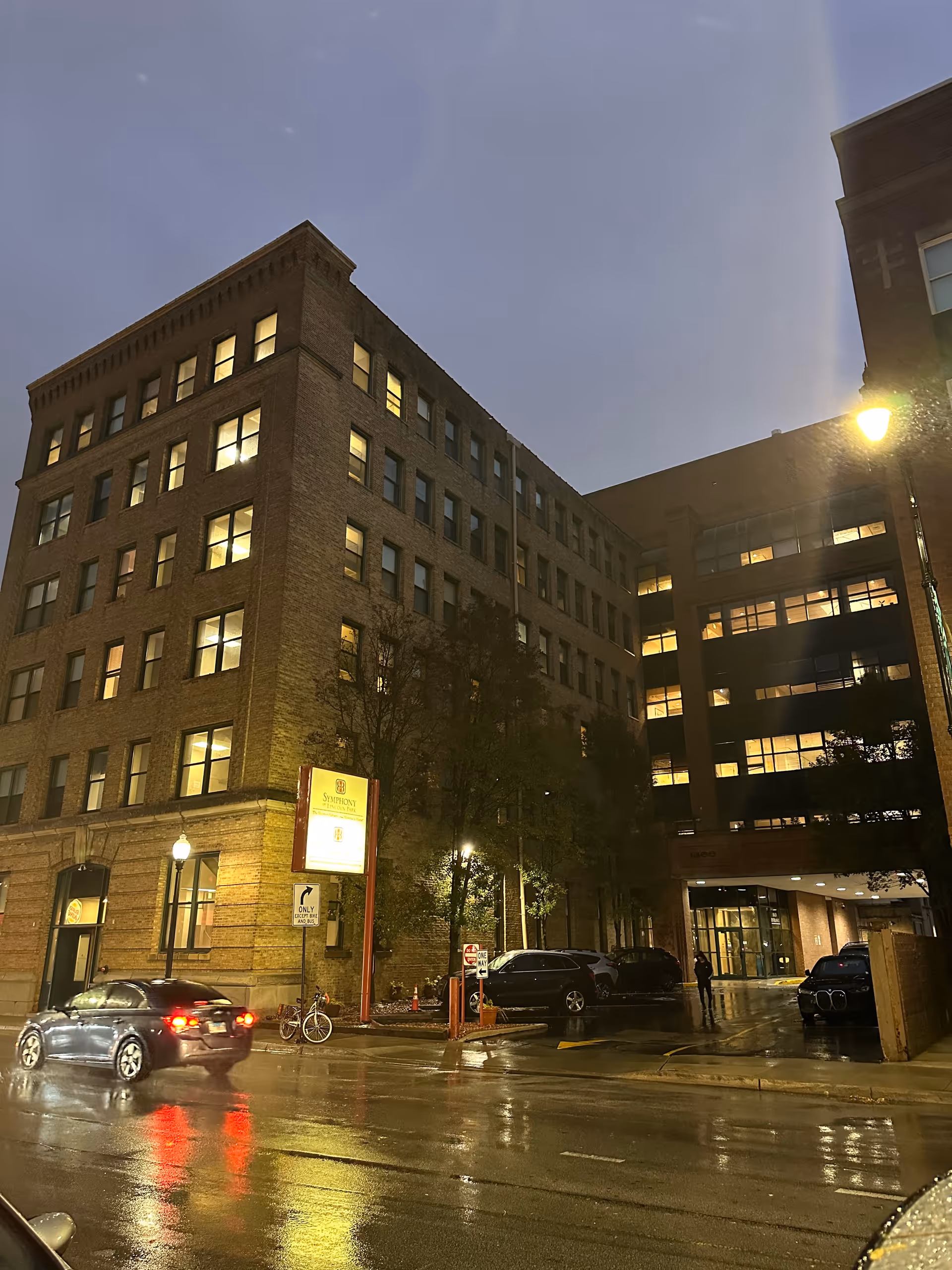 A nighttime street view of a multi-story brick building with many lit windows. There are cars parked along the street and in a parking area beside the building. The wet pavement reflects the streetlights and car lights. A sign near the corner reads 'Symphony of Lincoln Park'.
