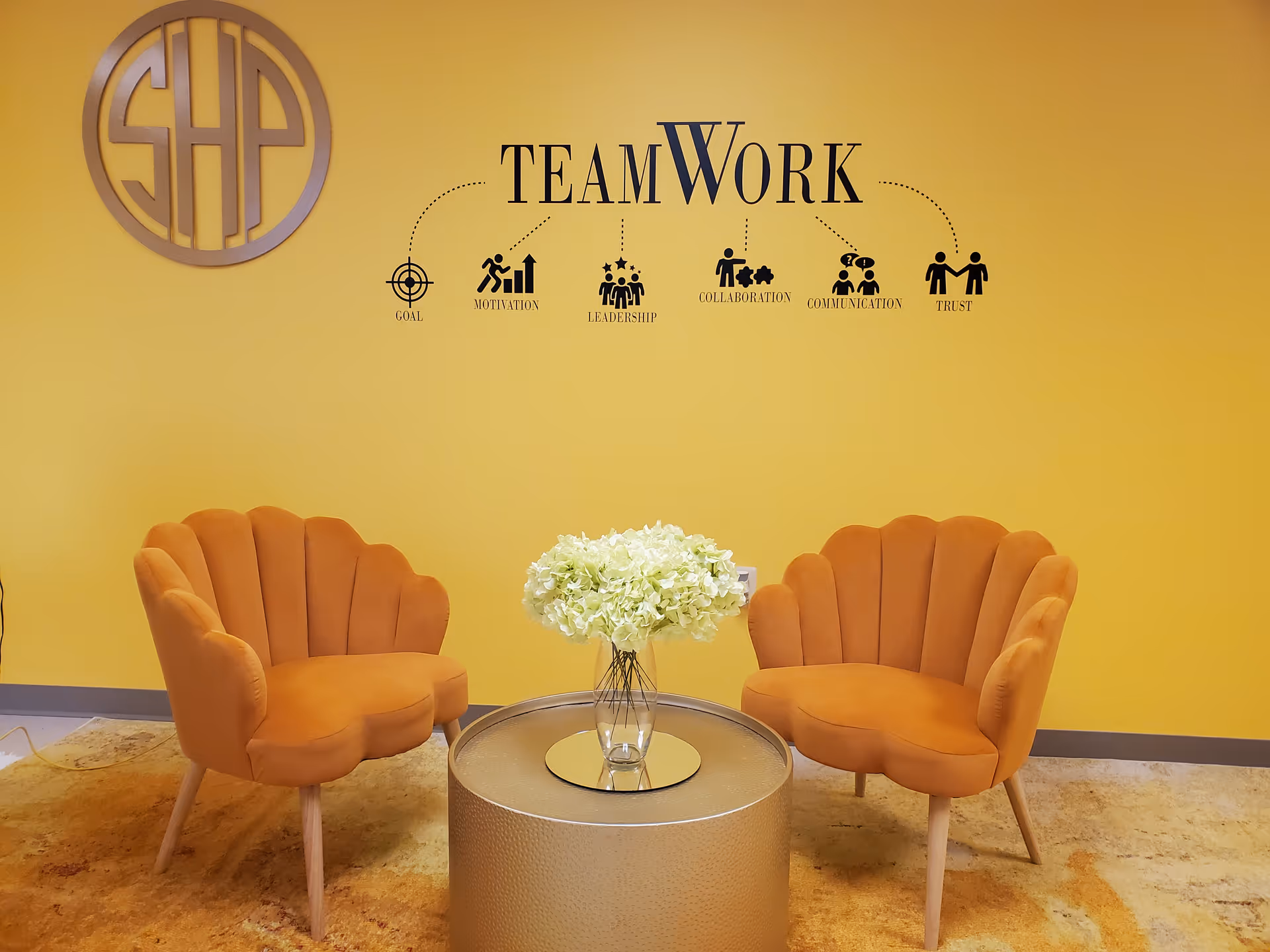 Two orange lounge chairs face a glass-topped table with flowers in a bright yellow lobby.