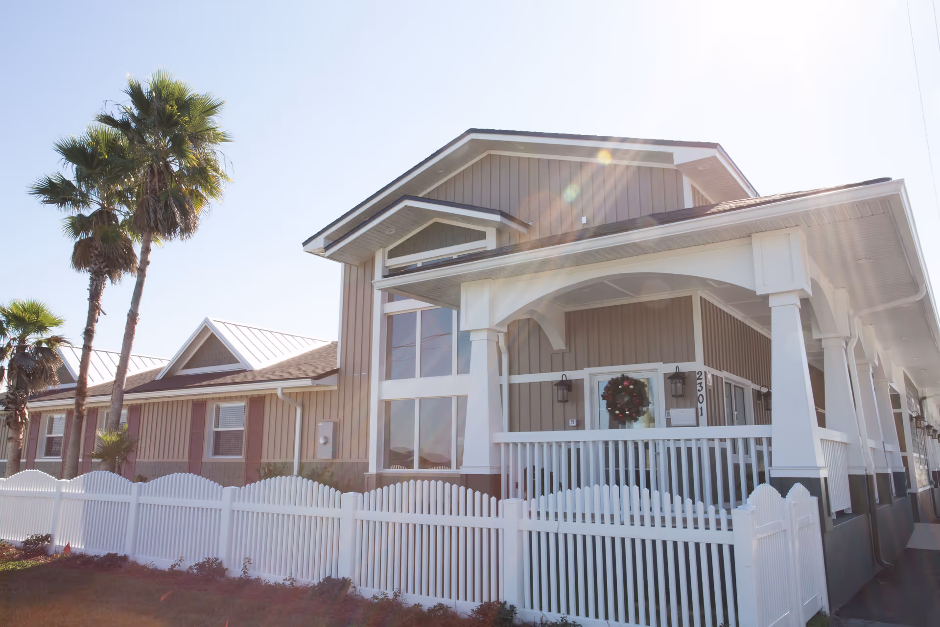 Exterior view of a single-story building with beige siding, white trim, and a white picket fence. The building has a covered porch decorated with a holiday wreath on the door. There are palm trees in the background and sunlight creating lens flare in the image.