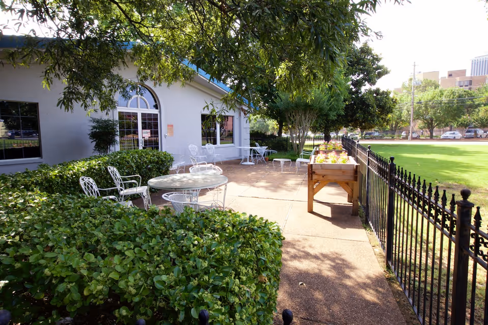 A view of the outdoor patio area at AHC Bright Glade, featuring white metal chairs and tables surrounded by lush greenery and a flower bed, with a building entrance visible in the background.