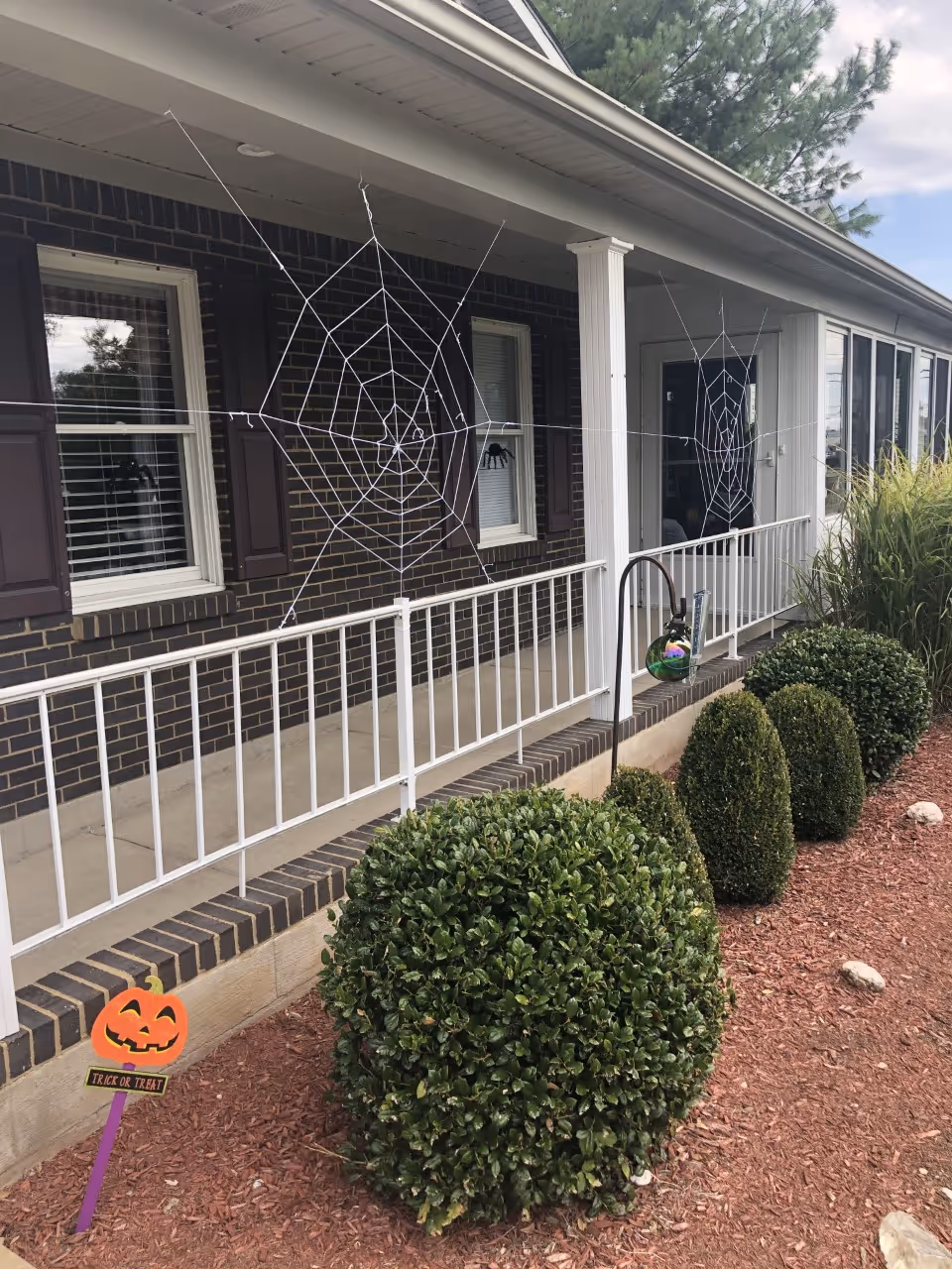 Front porch of a brick building decorated with two large spider webs and a small pumpkin sign that says 'Trick or Treat'. There are neatly trimmed bushes and a garden area with mulch in front of the porch, which has white railings and columns.