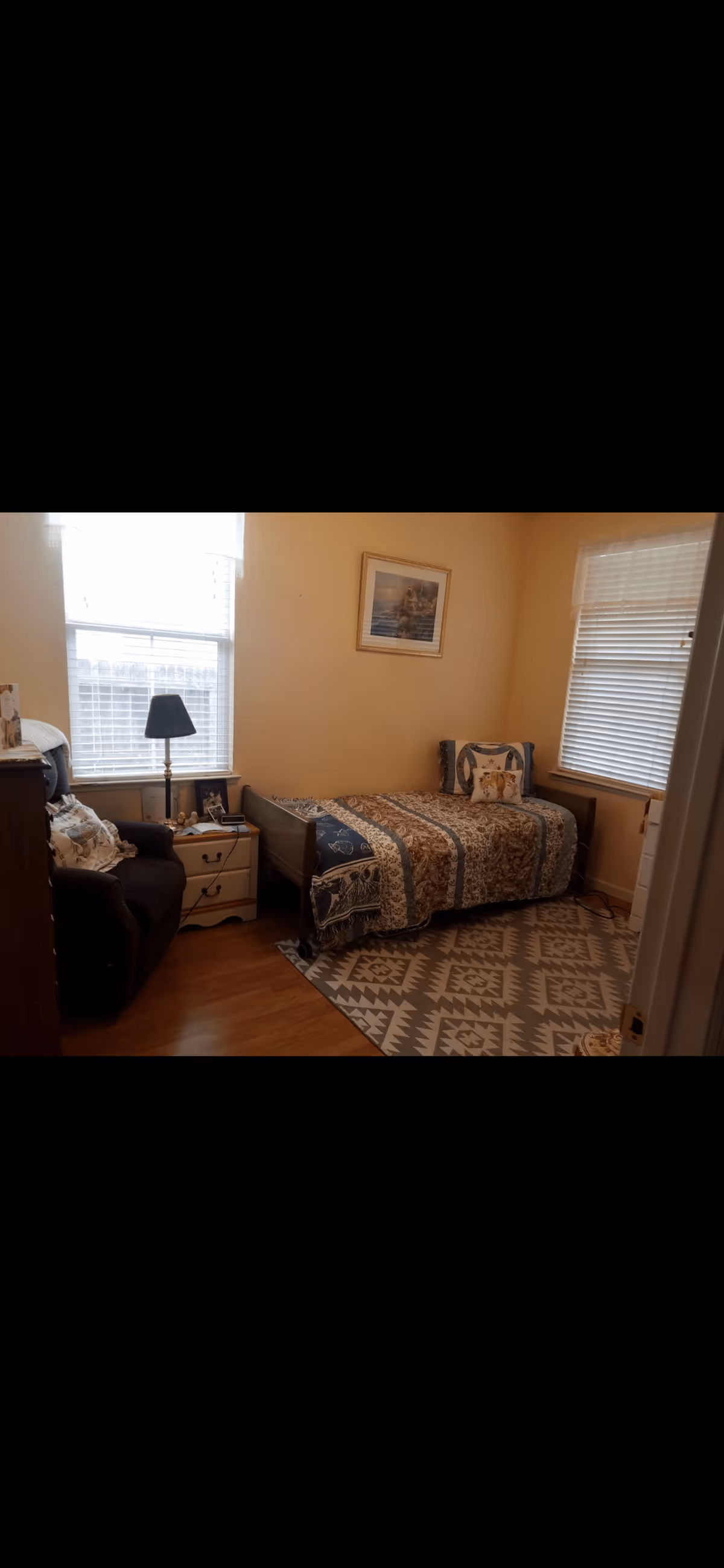 A small bedroom with a single bed covered in a patterned quilt and pillows. There is a nightstand with a lamp and some items next to the bed. A dark armchair is positioned near the window with white blinds. The room has light-colored walls, a framed picture hanging above the bed, and a patterned rug on the wooden floor.