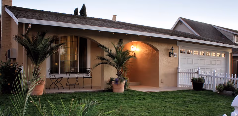Exterior view of a single-story house with a covered porch featuring two potted palm plants and a small table with two chairs. The house has a beige stucco finish, a shingled roof, a white garage door, and a white picket fence. The lawn in front is green and well-maintained, and a porch light is turned on near the entrance.