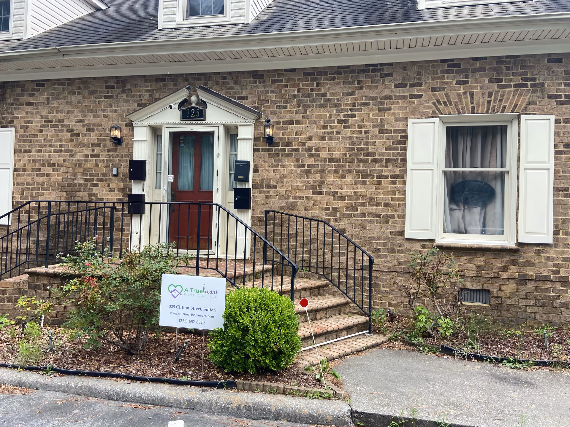 Brick facility entrance with red door, stairs, railing, and A Trueheart Home Care sign.