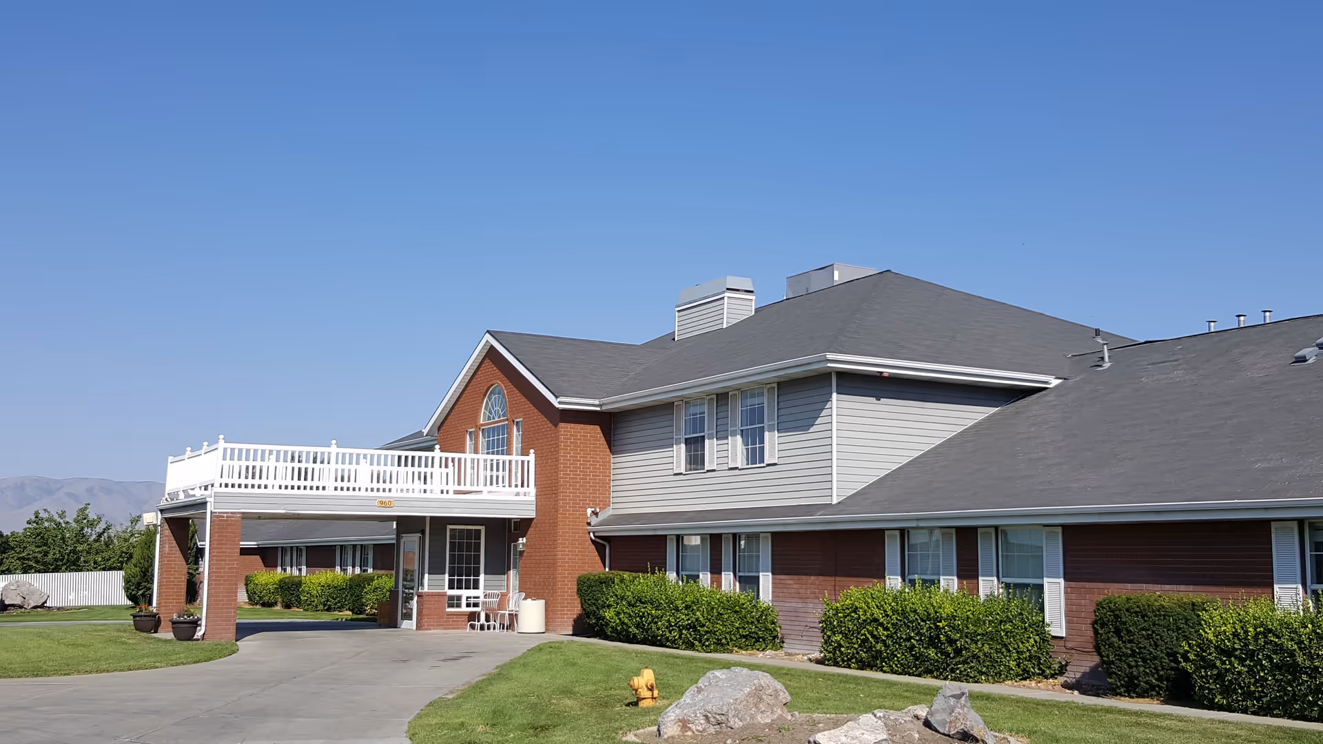 Front exterior of a two-story red brick and gray senior living building with a balcony, driveway, and landscaped lawn under a clear blue sky.