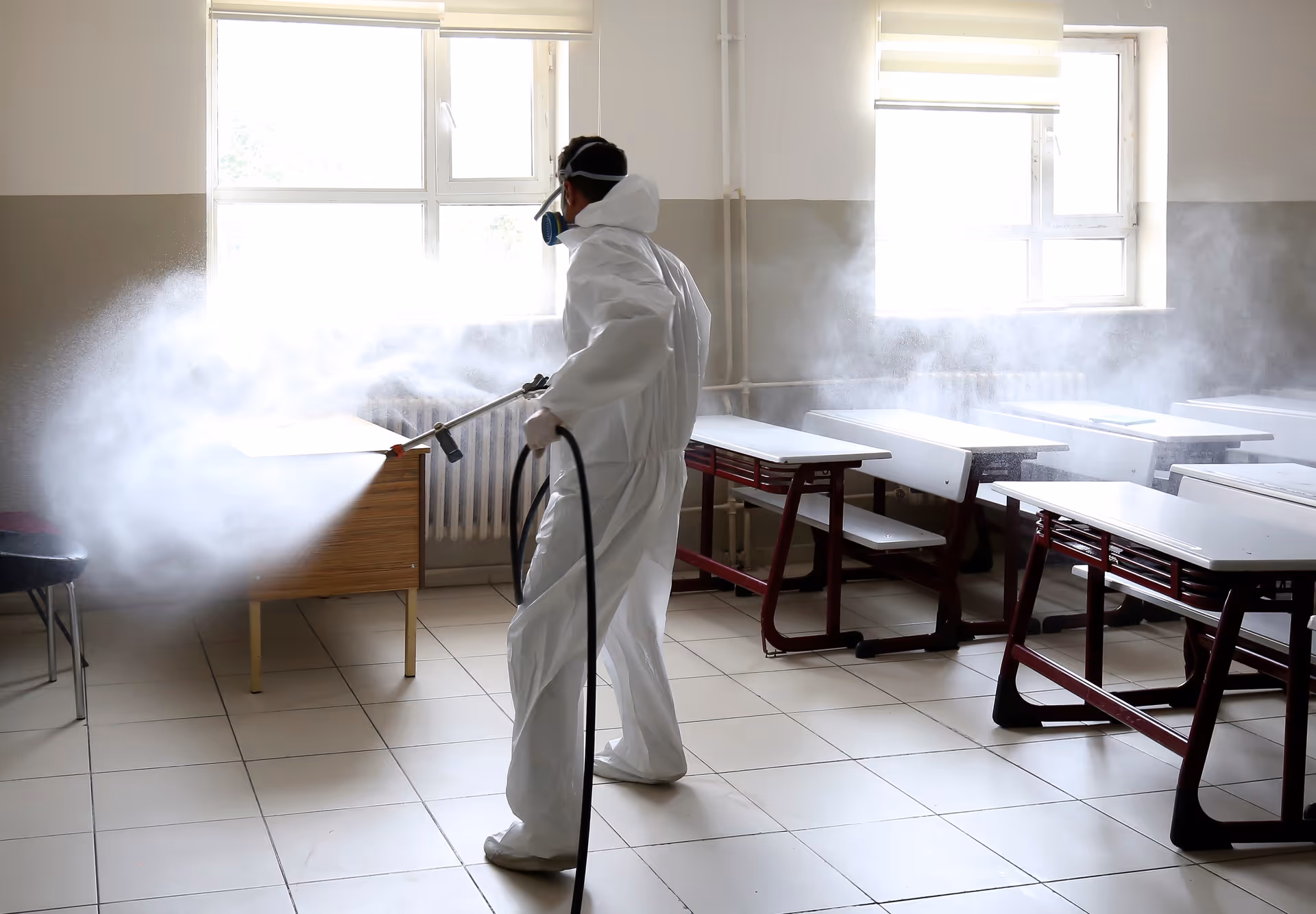 A worker in protective PPE disinfects a classroom with a sprayer, creating a mist.