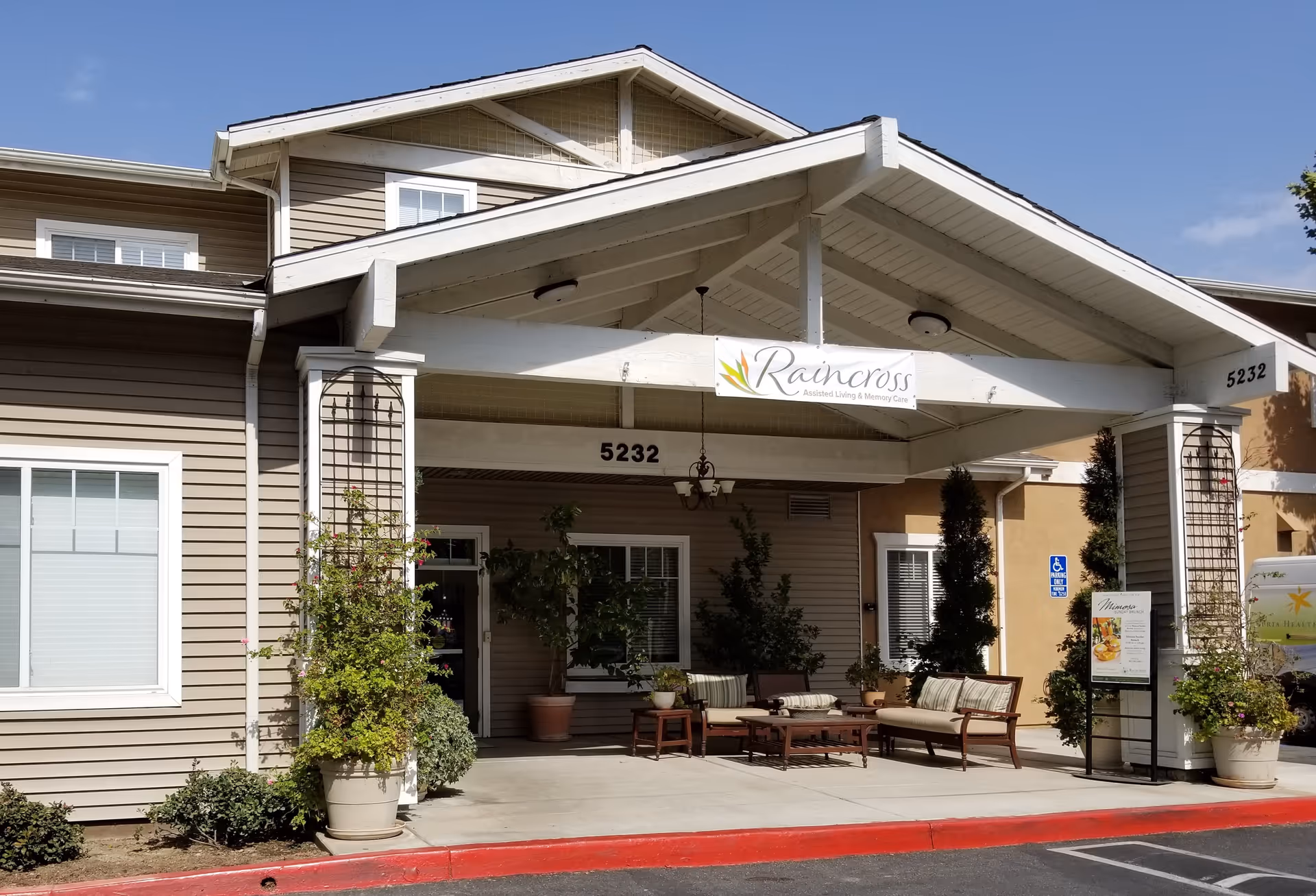 Exterior view of a senior living facility entrance with outdoor seating and greenery.