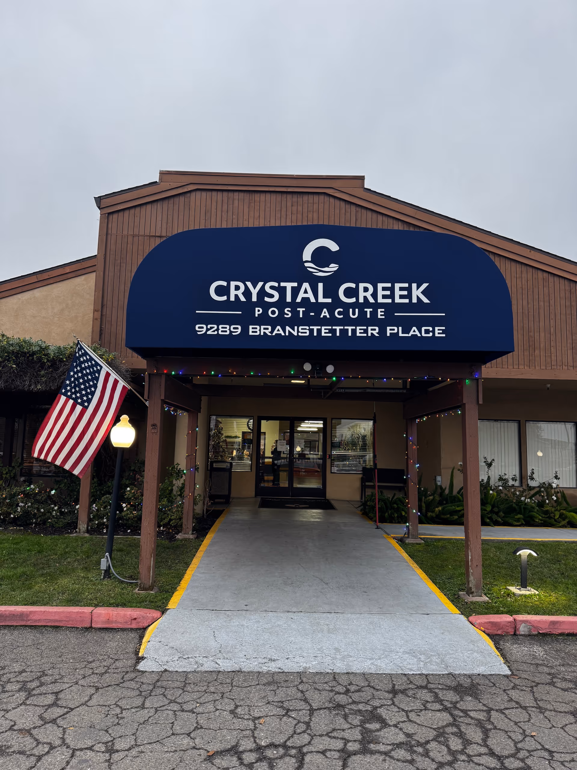 Front entrance of a senior care facility with a blue Crystal Creek Post-Acute sign and flag.