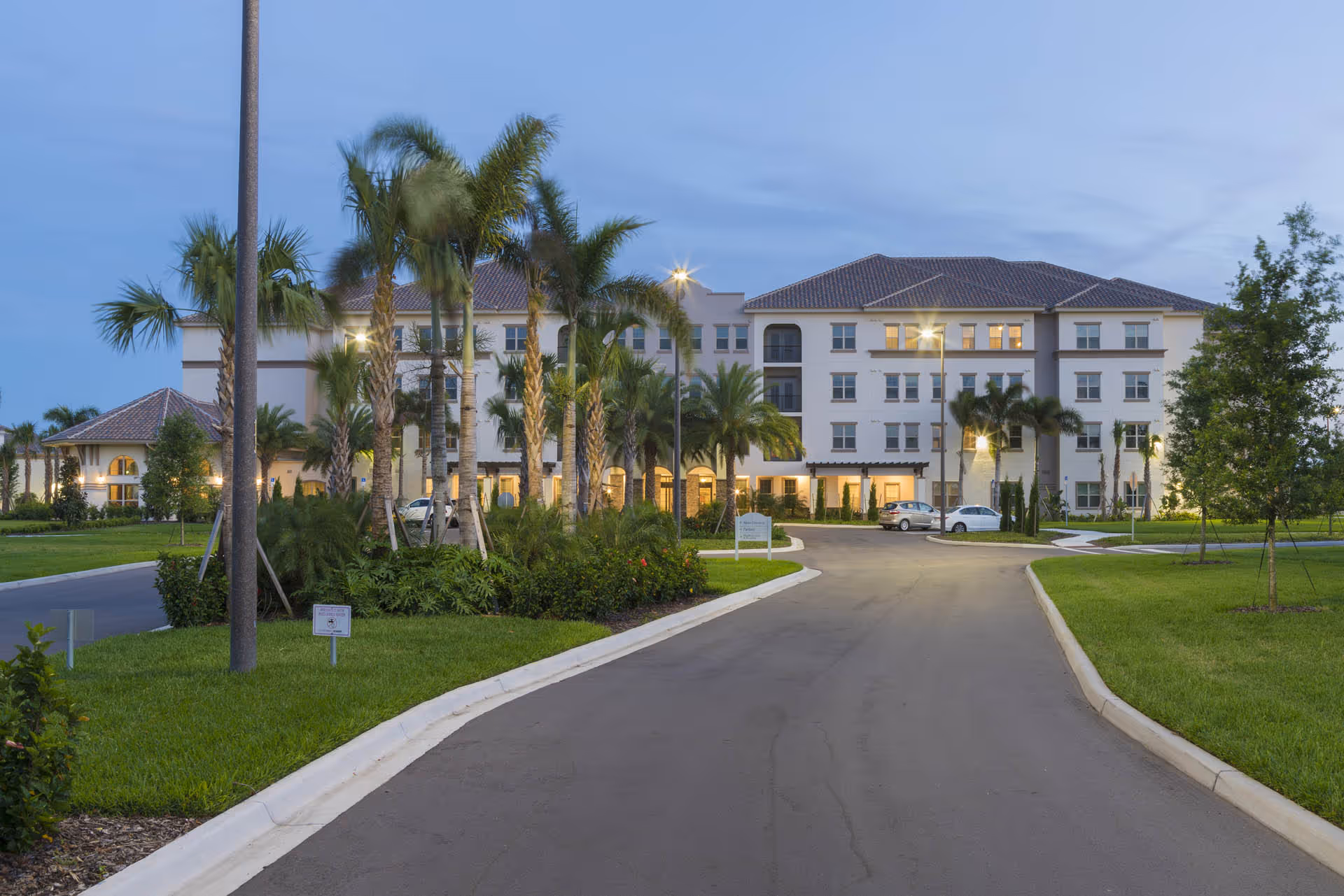 Front exterior of a multi-story senior living building with a driveway, palm trees, and evening lights.
