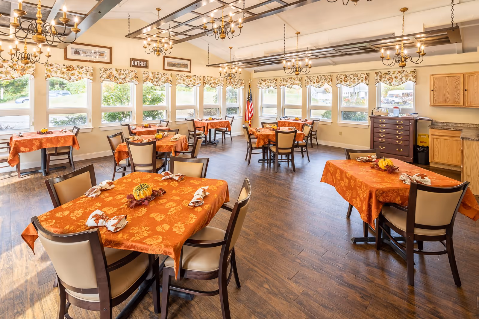 A bright dining room with multiple tables covered in orange tablecloths with autumn leaf patterns. Each table is set with napkins and small decorative centerpieces featuring pumpkins and fall leaves. The room has large windows with floral valances, wooden flooring, chandeliers hanging from the ceiling, and an American flag in the corner. There is a wooden cabinet and countertop along one wall.