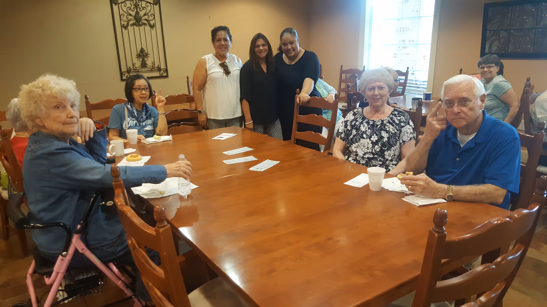 Senior residents and caregivers sit and stand around a large wooden dining table.