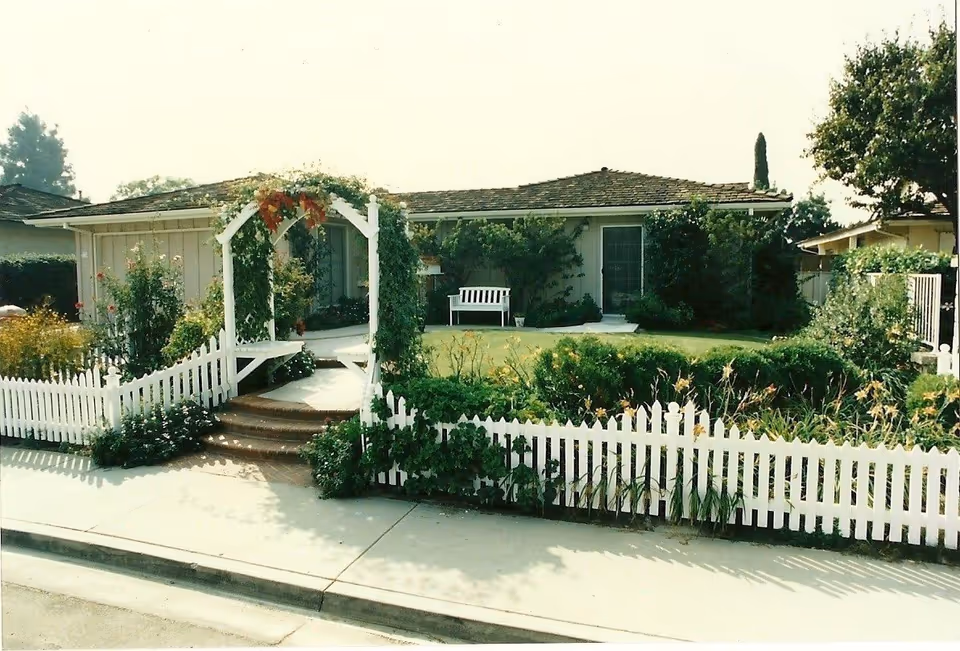 Front view of a fenced yard with a white arch, garden, and bench.