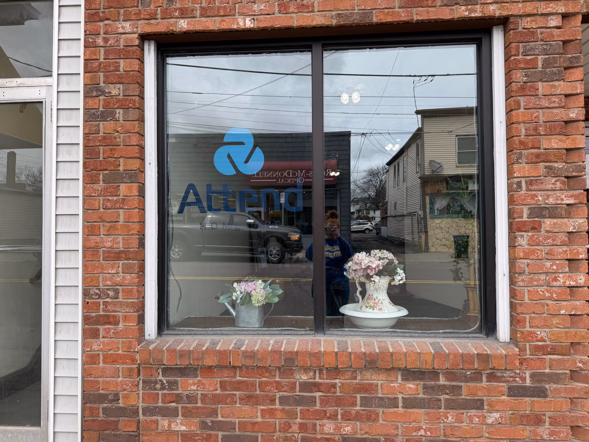 Brick storefront window with Attend Home Care logo and potted flowers, reflecting street scene.