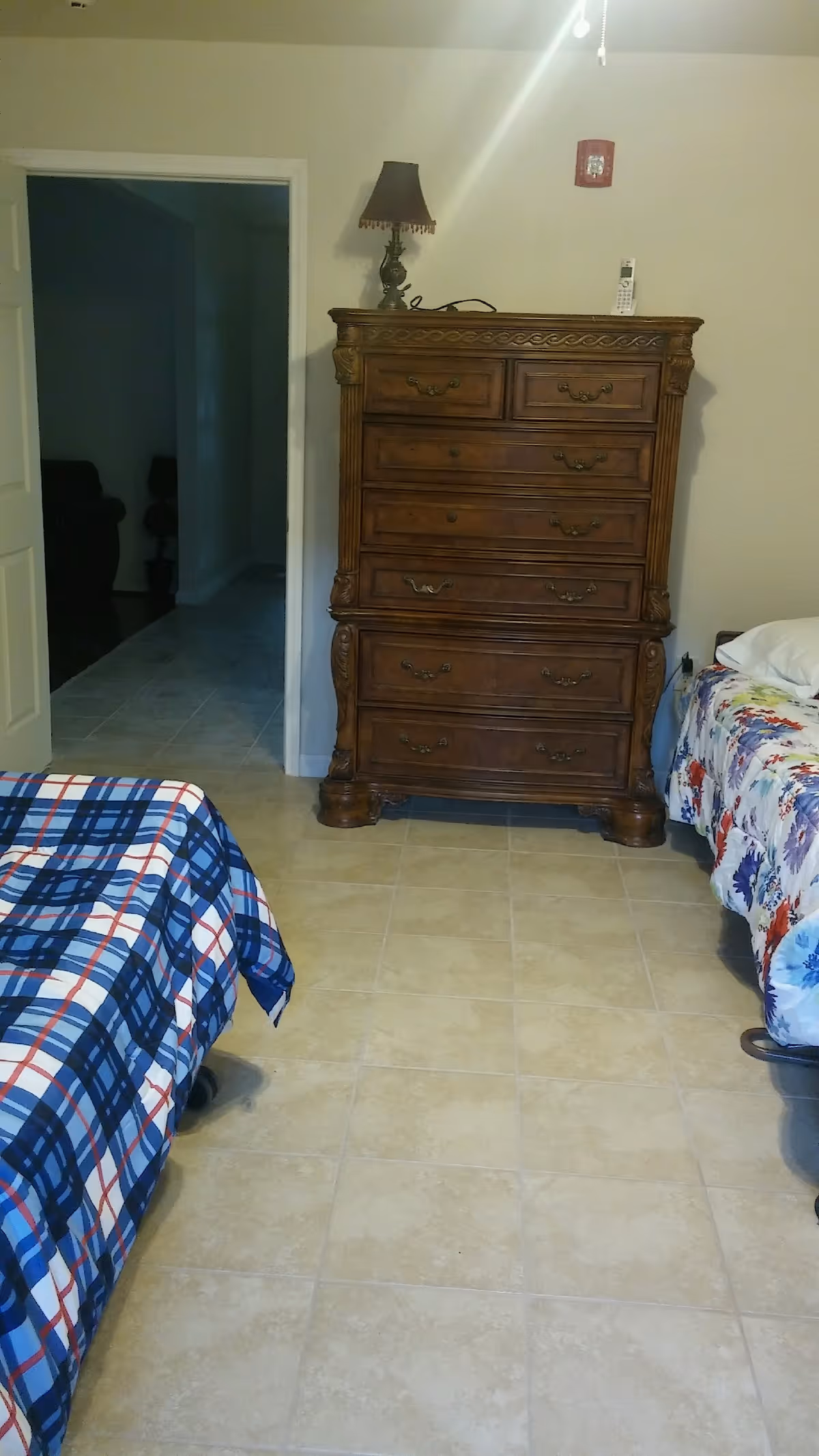 A bedroom with two beds, one with a blue and white plaid bedspread and the other with a floral bedspread. Between the beds is a large wooden dresser with multiple drawers, a decorative lamp, and a cordless phone on top. The room has tiled flooring and a doorway leading to a hallway.
