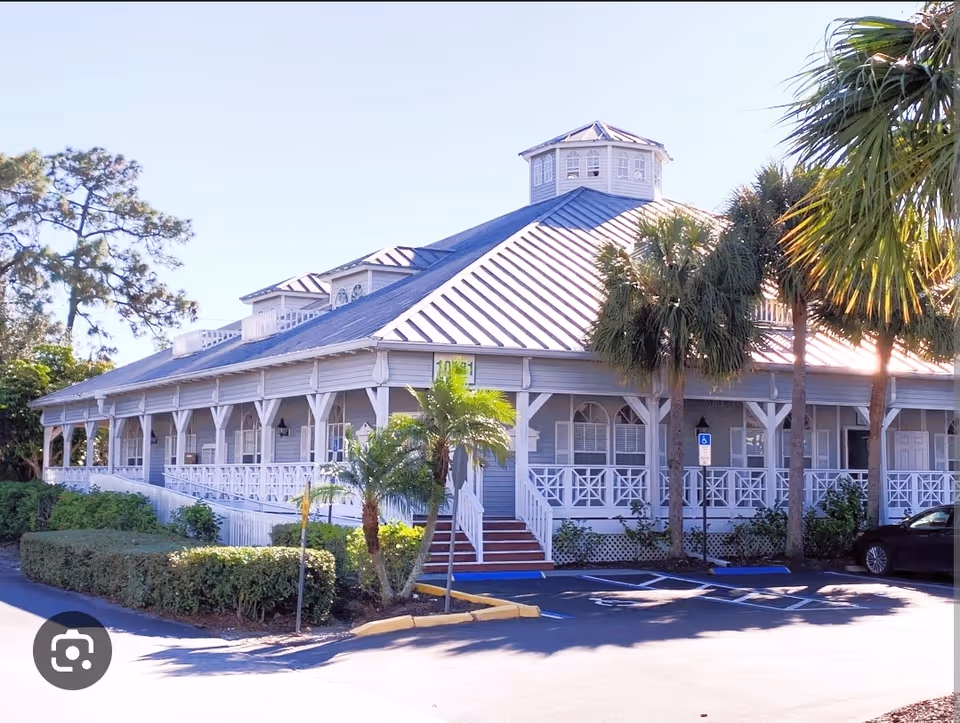Exterior view of a nursing facility with a wraparound porch and palm trees.