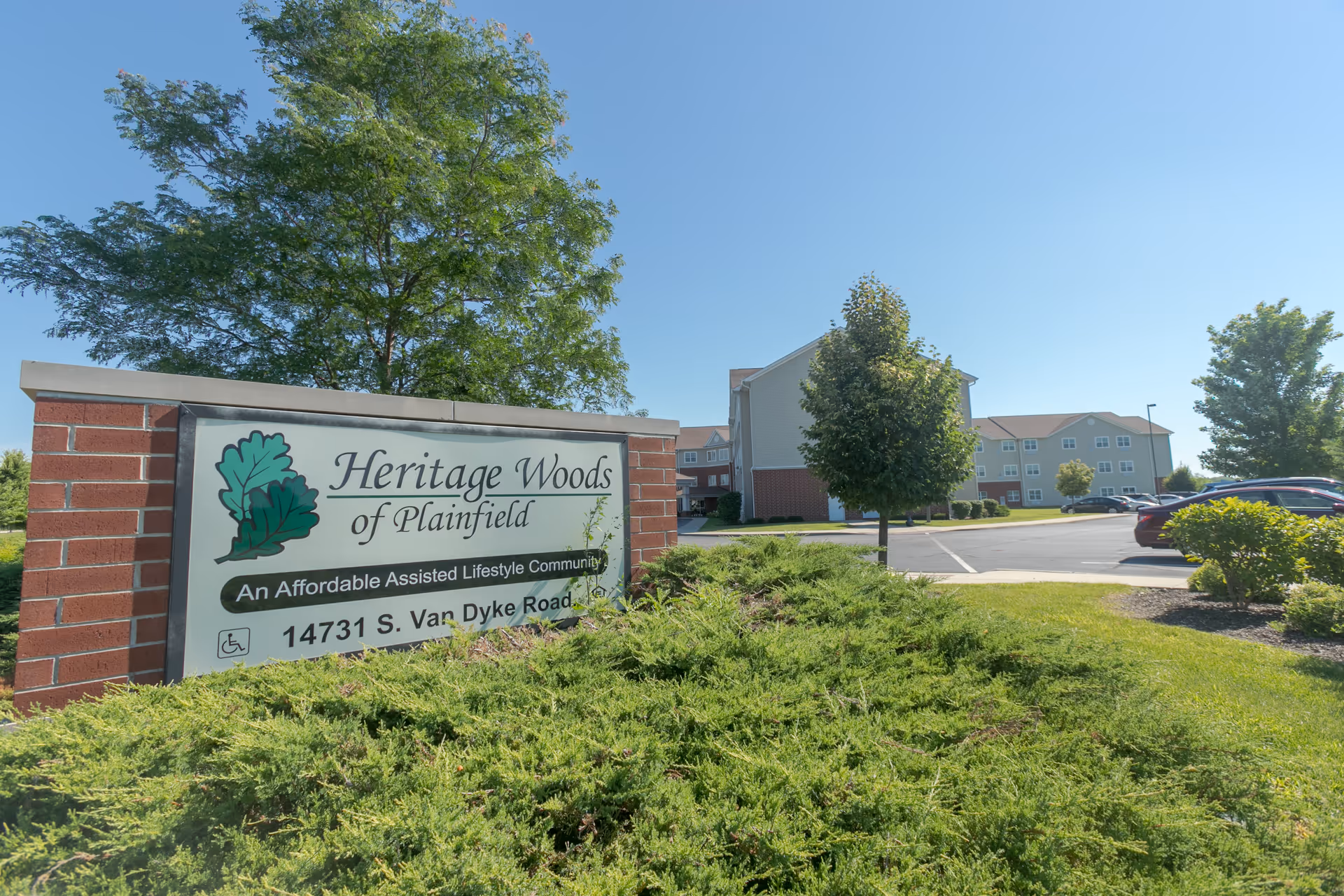 Entrance sign reading 'Heritage Woods of Plainfield' with the facility building, parking lot, and landscaping in the background.