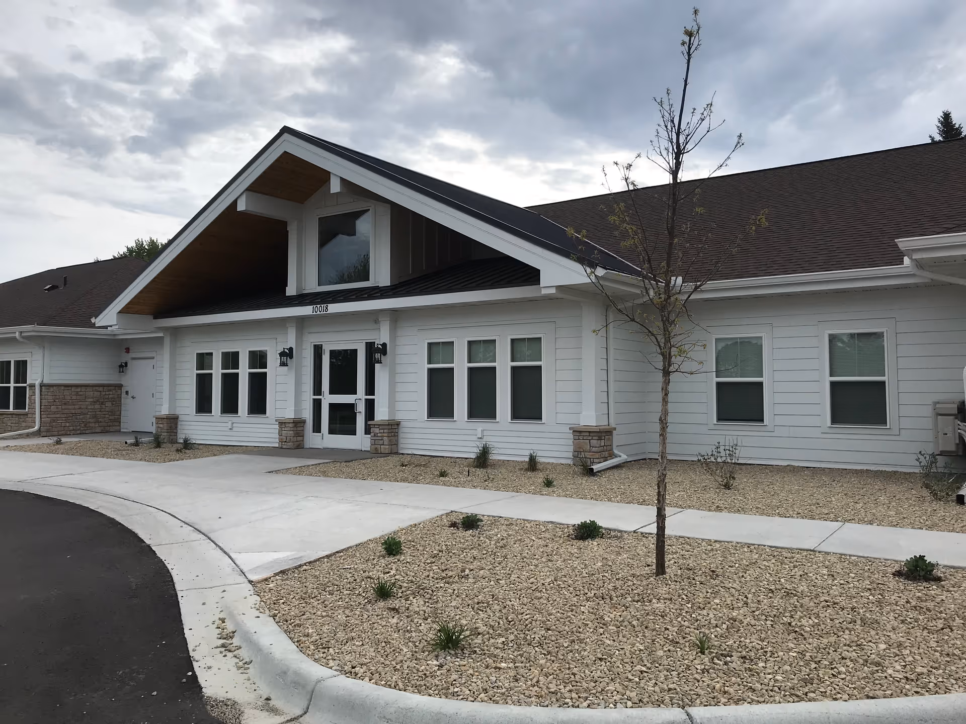 Front entrance of a single-story white building with a covered gabled entry, multiple windows, and gravel landscaping.