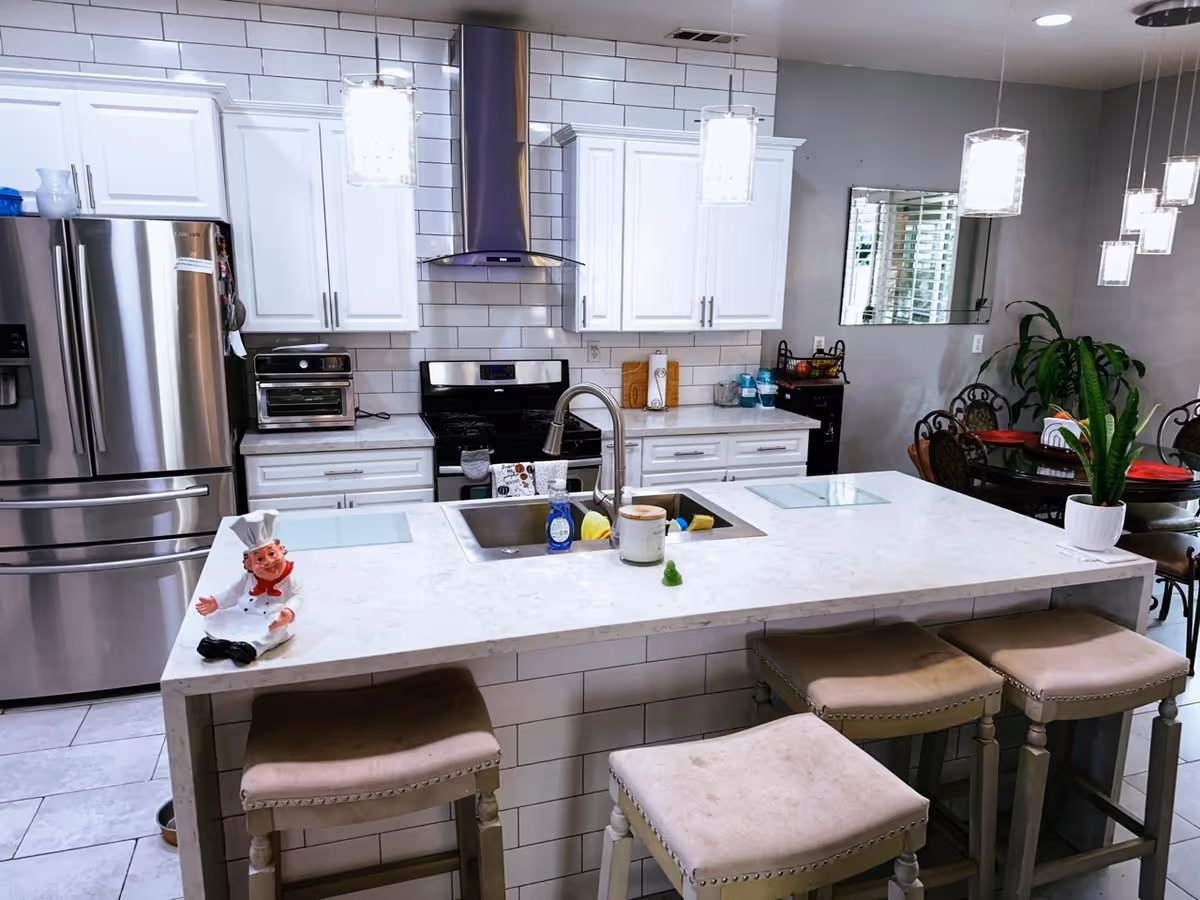 Modern white kitchen with island, sink, stainless appliances, pendant lights, and seating.