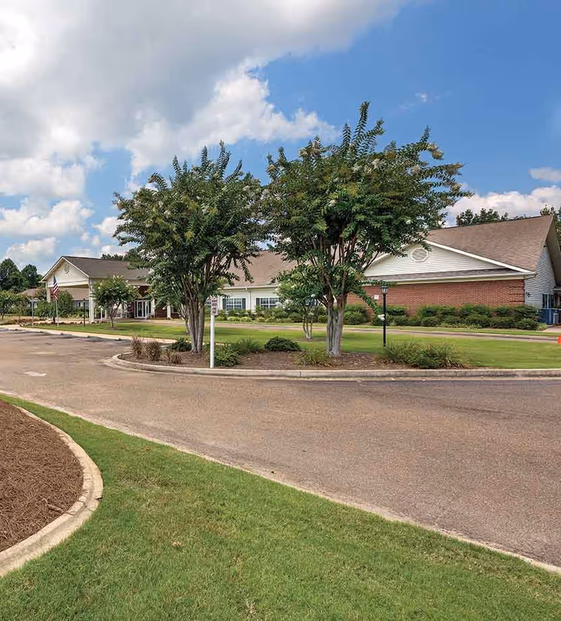 Exterior view of a single-story senior living facility building with a brick and white siding facade, surrounded by green grass, trees, and a paved driveway under a partly cloudy blue sky.