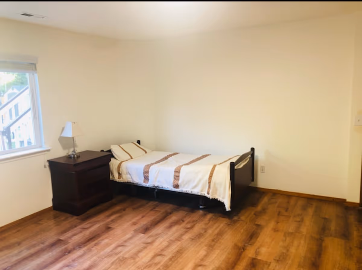A simple bedroom with a single bed, nightstand, lamp, and window in a calm room.